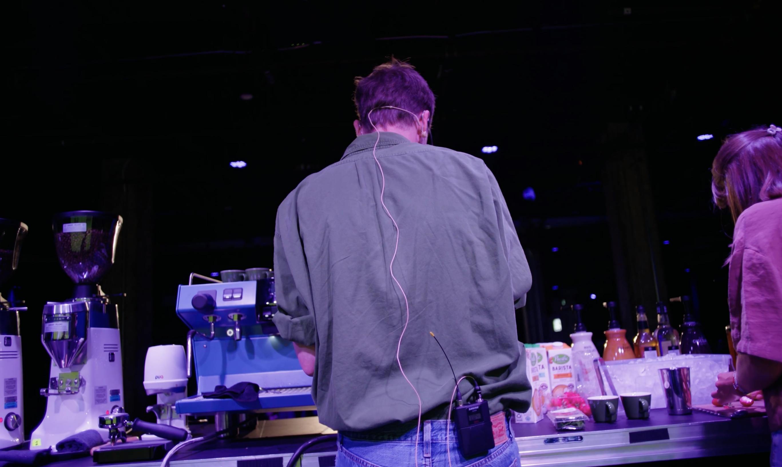 The Barista League back of man making coffee on stage with microphone lead down back