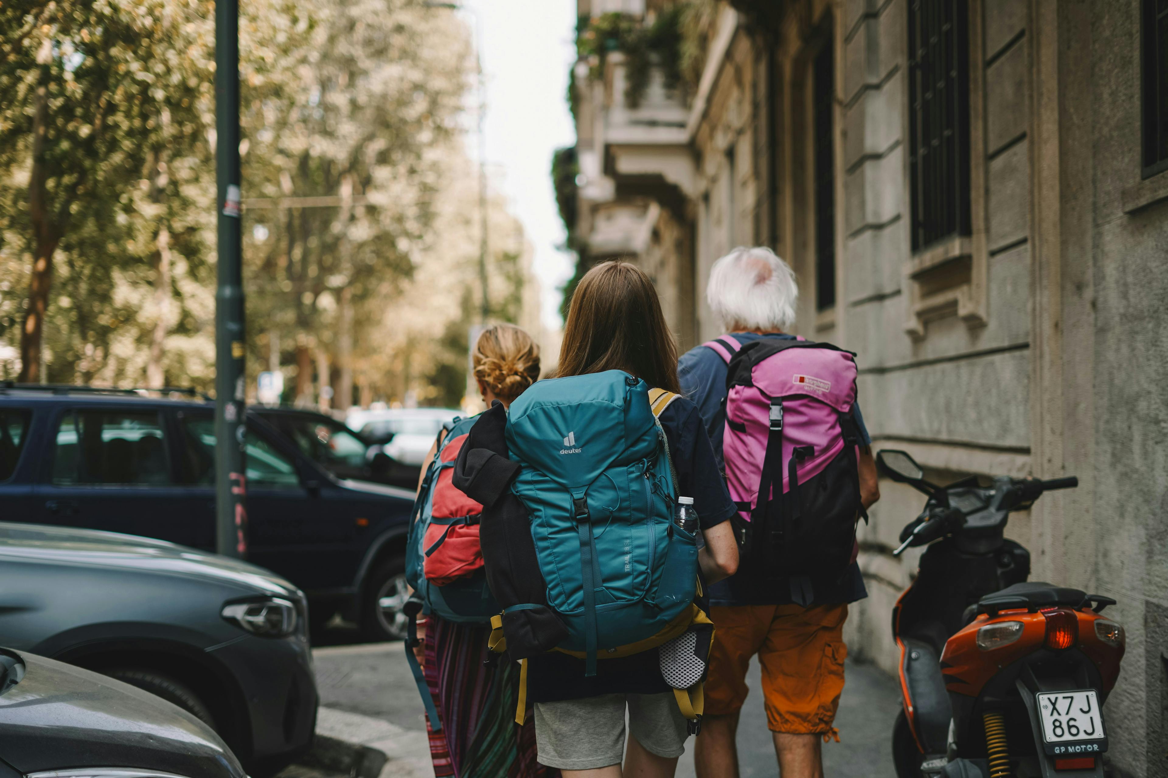 Three tourists walking down tree-lined city street with backpacks