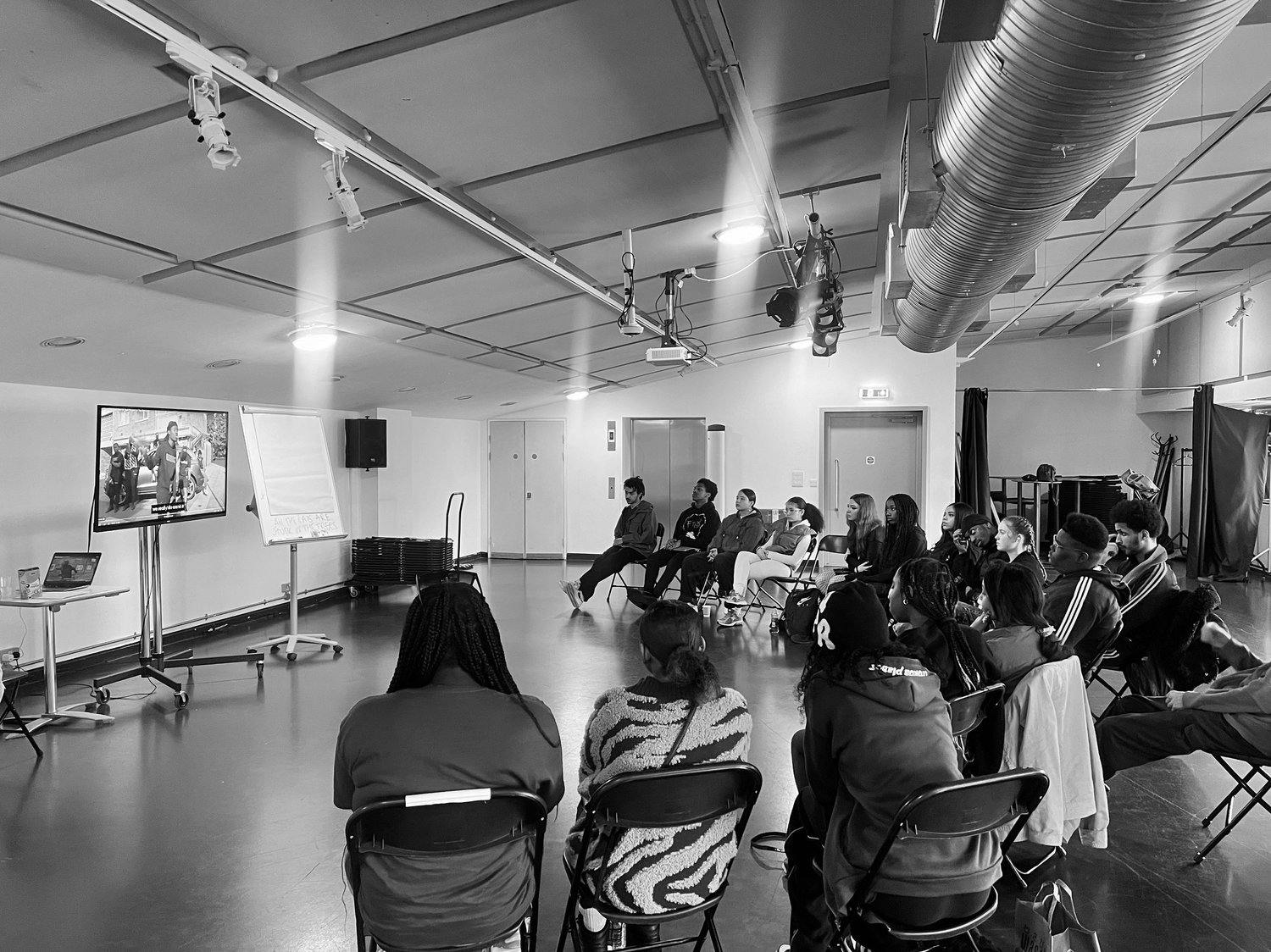 A black and white photo of a group of people sitting on chairs watching a TV screen in a conference room