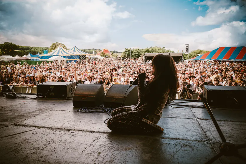 woman kneeling on stage singing into mic in front of large festival crowd