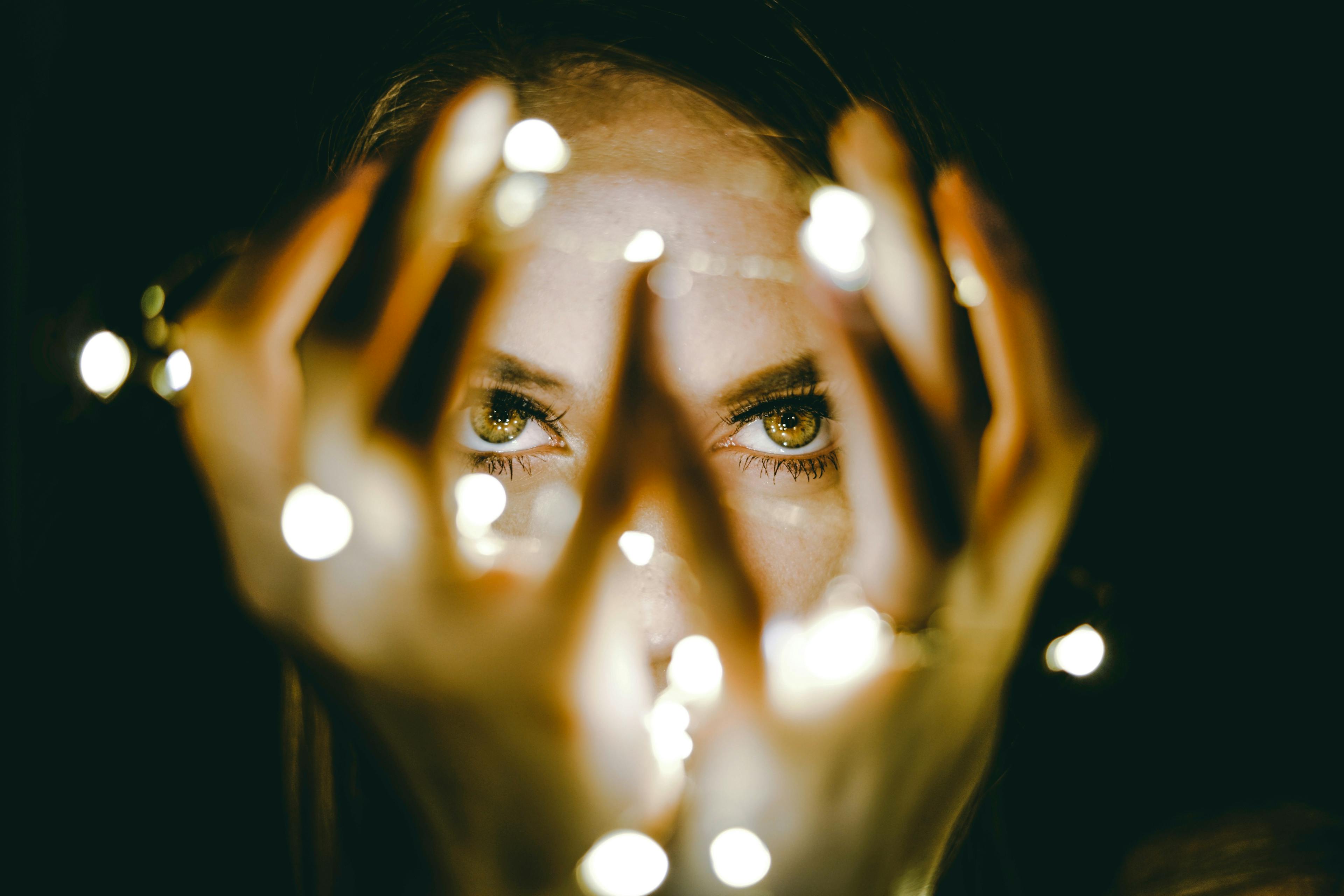 Woman's face visible through held up hands with glowing fairy lights